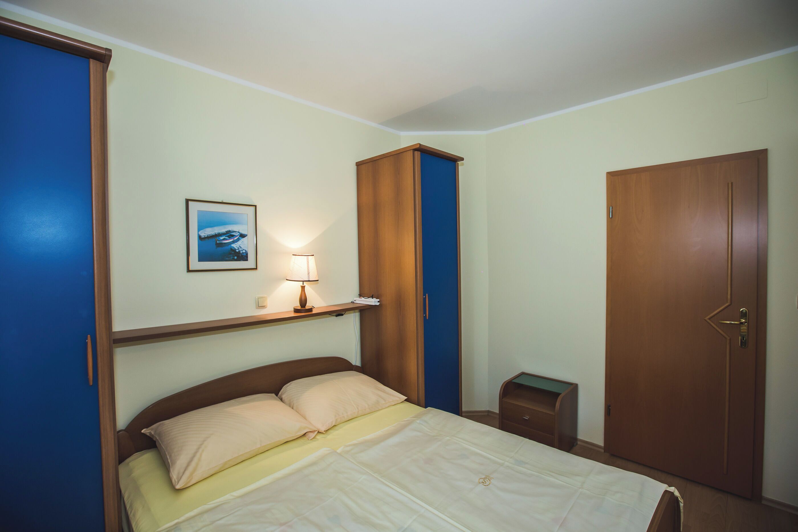 A bedroom with a bed, a nightstand, a wardrobe, and a framed picture on the wall.