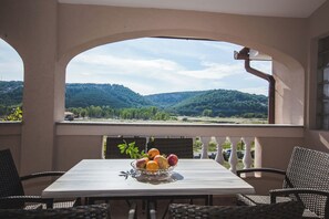 A balcony with a table and chairs, a bowl of fruit, and a view of green hills. - Apartments Anamarija S (Lopar)