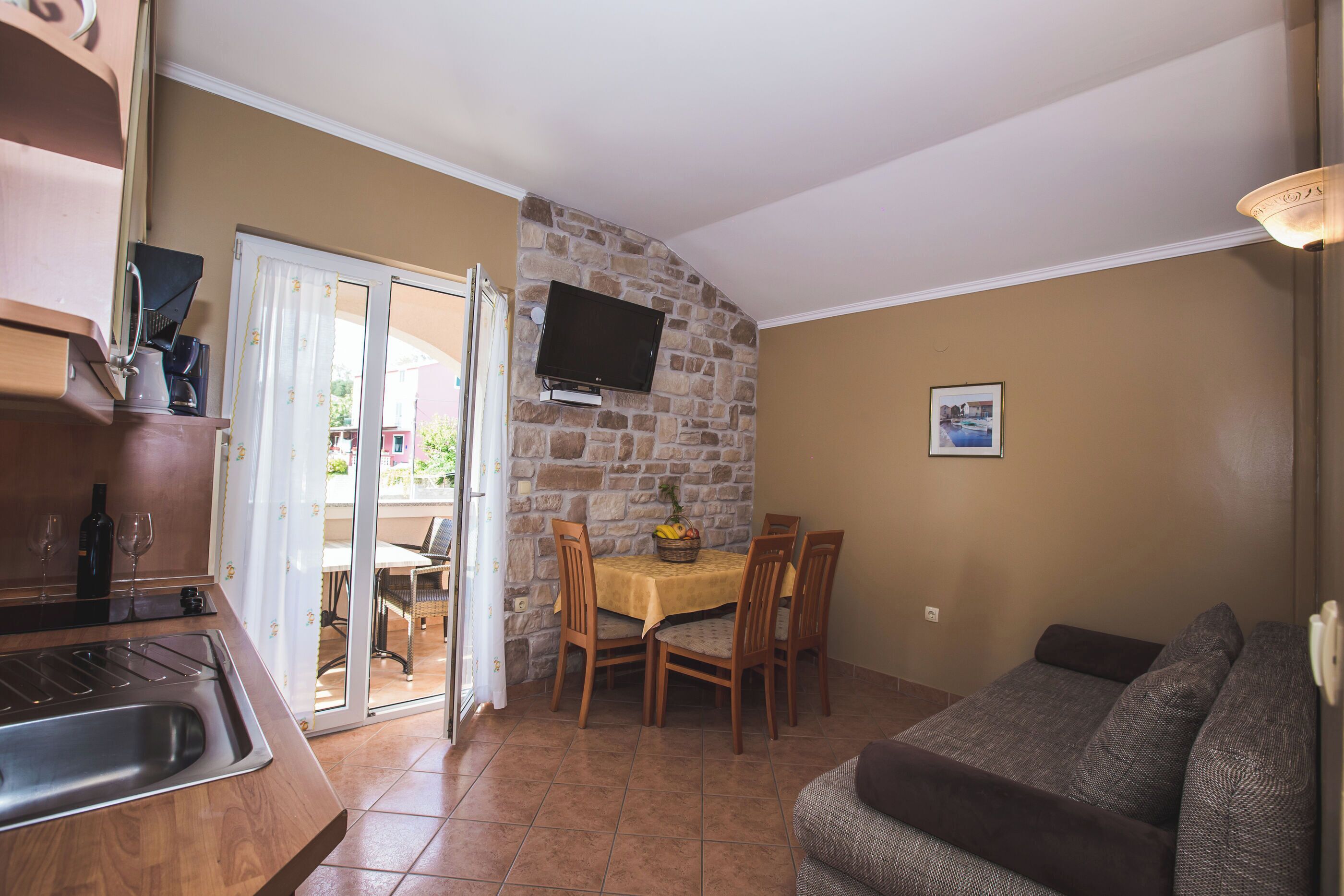 Espai vital compacte amb zona de cuina, taula de dinar amb cadires, televisió de pantalla plana penjada a una paret de pedra i sofà.