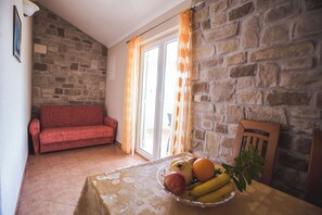 A room with a stone wall, a red sofa, a dining table with a fruit bowl, and a wooden chair. - Apartments Anamarija S (Lopar)