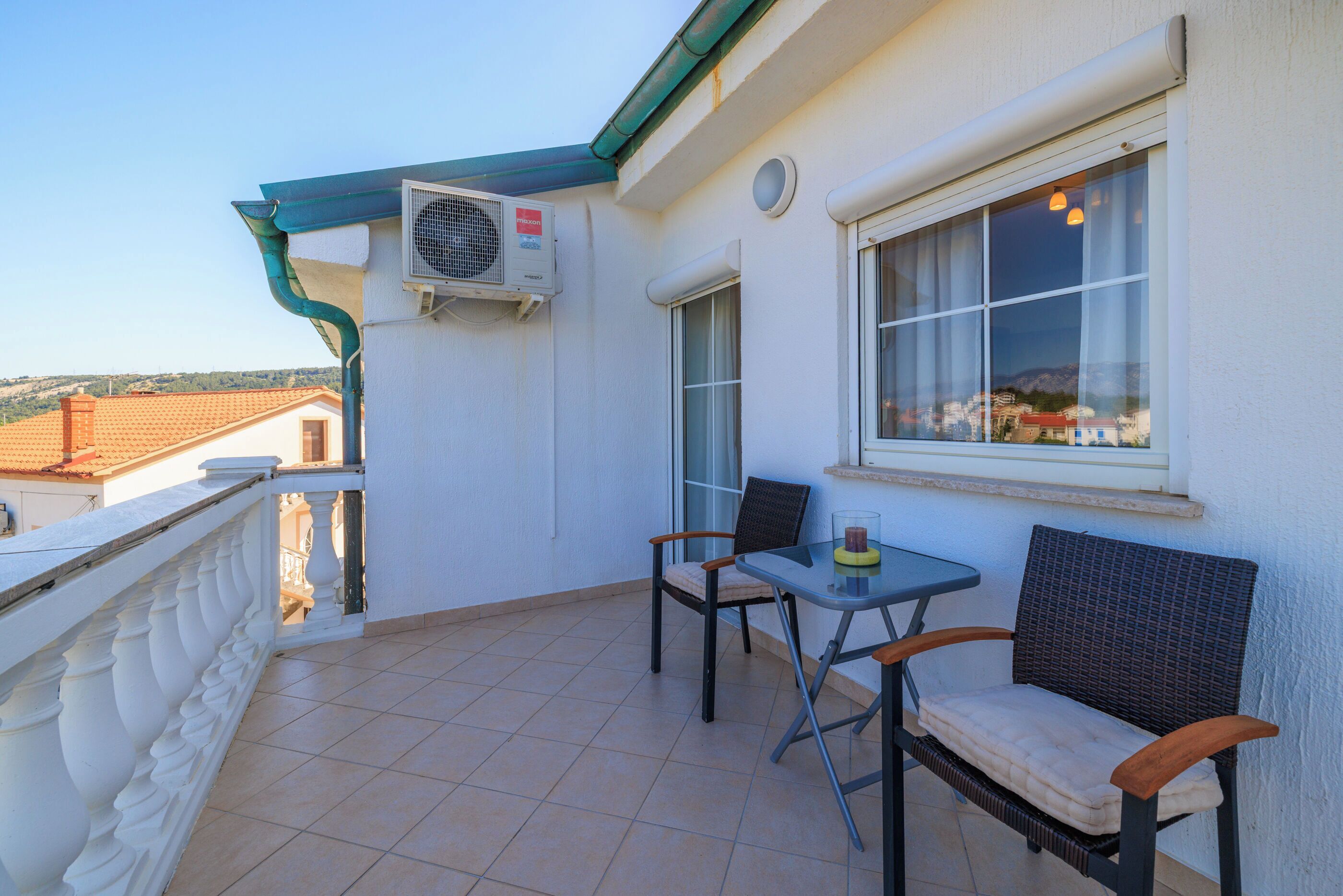 A balcony with a table and two chairs, an air conditioning unit, and a window with curtains.