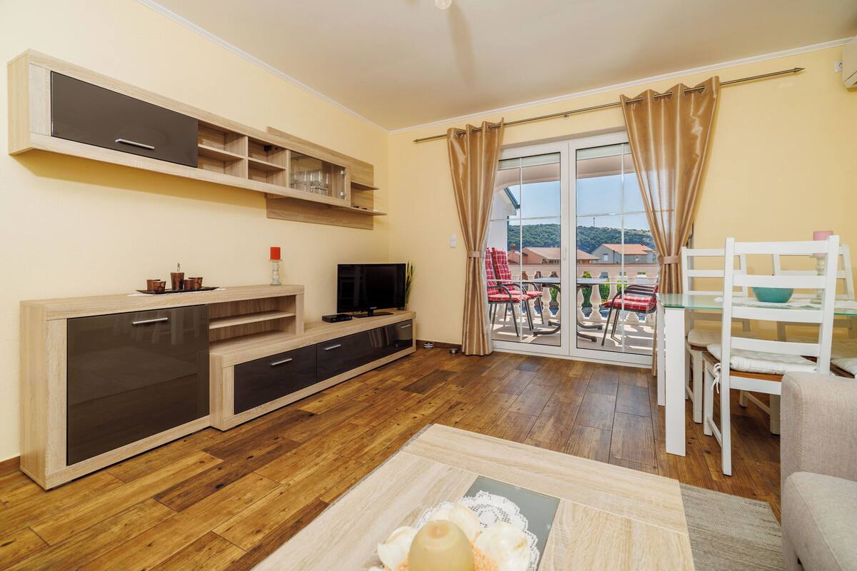 A modern living room with a wooden floor, a built-in entertainment unit, a dining area with a glass table and chairs, and a view through a sliding glass door.