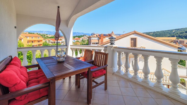 Un balcón con una mesa de madera y cojines rojos, con vistas a un paisaje urbano que incluye edificios y montañas.