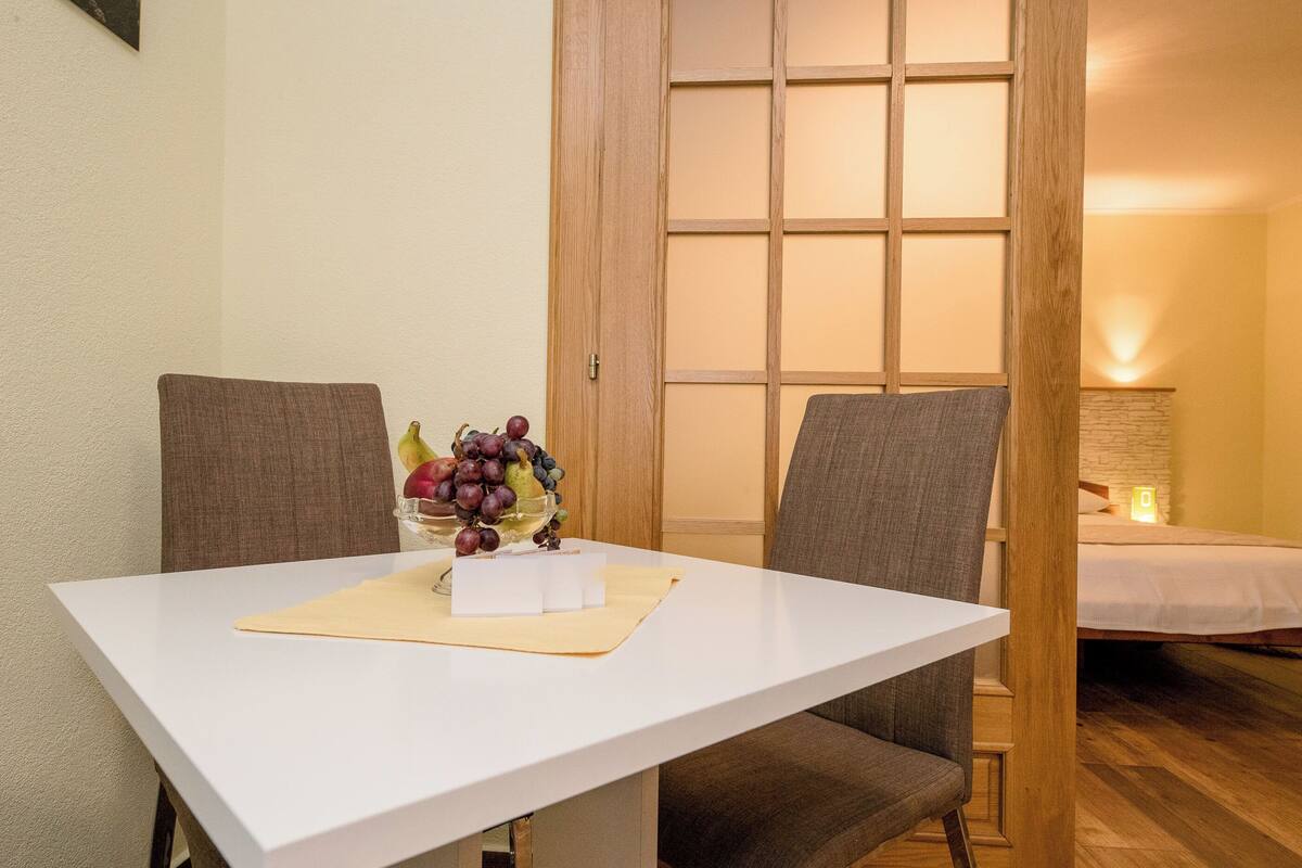 A dining table with a fruit bowl, two chairs, and a bed in the background.