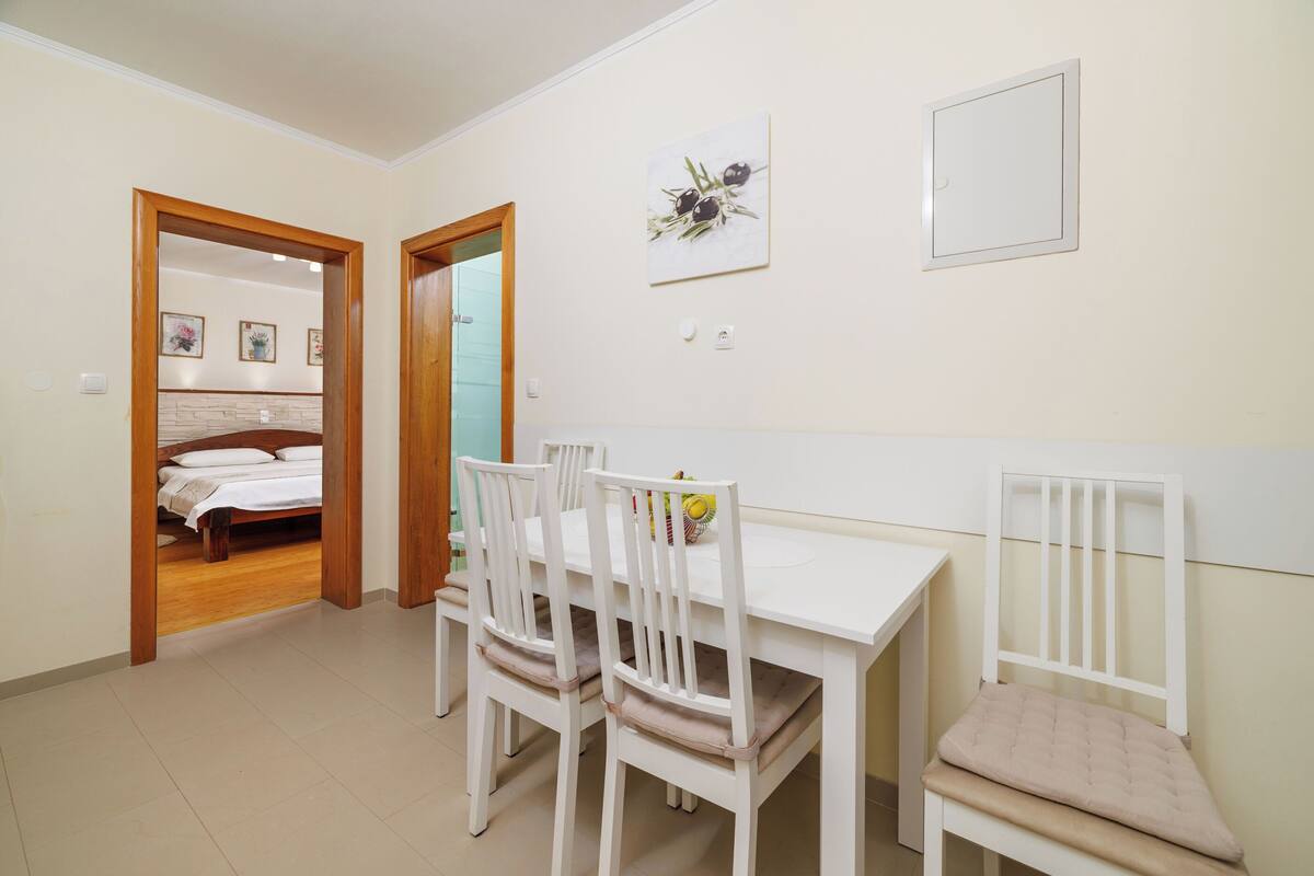 A dining area with a white table and chairs, a bed in the adjacent room, and a wall-mounted light switch.