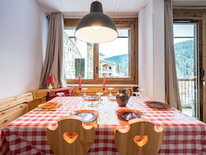 A dining area with a checkered tablecloth, wooden chairs with heart cutouts, a candle, and a view of a mountain landscape through large windows. - T3 Comfort Courchevel Moriond - 6 pers, Ski-in/out, Parking (Courchevel)