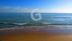 Une plage aux eaux calmes, un ciel dégagé et quelques voiliers à l’horizon.