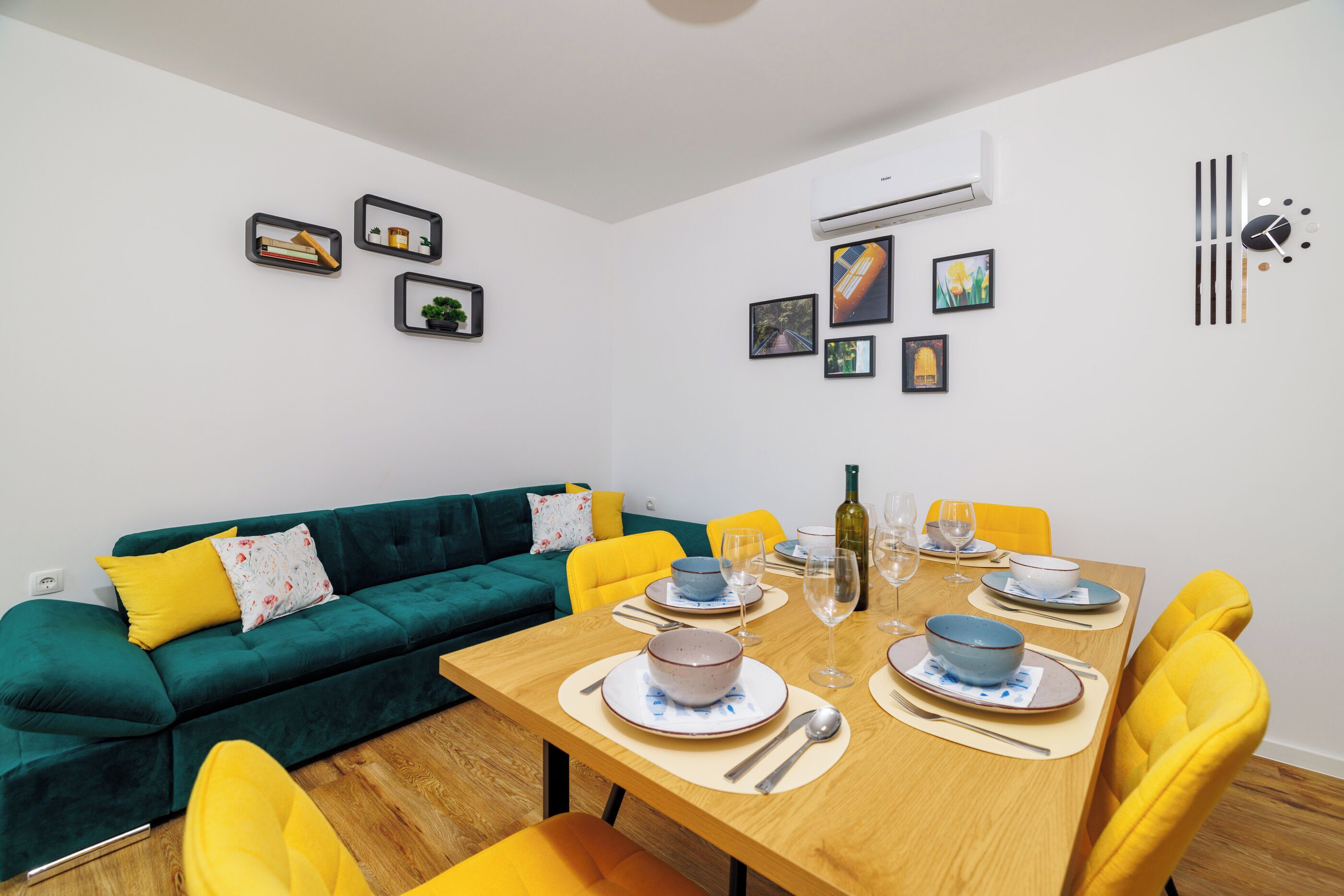 Un coin repas avec une table en bois dressée pour quatre personnes, entourée de chaises jaunes. Un canapé vert avec des coussins jaunes est adossé au mur, et des tableaux encadrés sont accrochés au mur au-dessus du canapé.