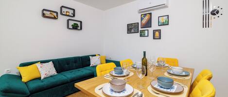 Un coin repas avec une table en bois dressée pour quatre personnes, entourée de chaises jaunes. Un canapé vert avec des coussins jaunes est adossé au mur, et des tableaux encadrés sont accrochés au mur au-dessus du canapé.