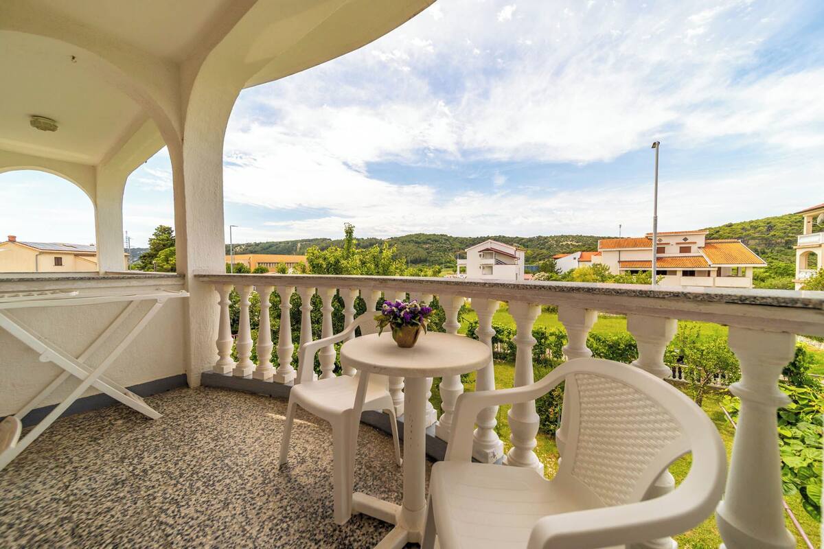 A balcony with white chairs and a small table, a view of greenery and buildings in the background.