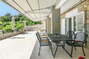 Eine Terrassenanlage mit Tisch und Stühlen, einem überdachten Bereich, Steinmauern und viel Grün im Hintergrund.