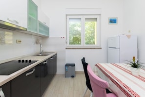 Cocina compacta con lavabo, estufa y armarios, un pequeño cubo de basura, una ventana con vistas a la vegetación y una zona de comedor con mesa y sillas.