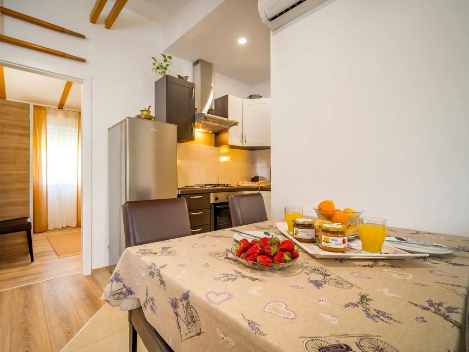 A dining area with a table set for two, a bowl of strawberries, a glass of orange juice, and a jar of jam.