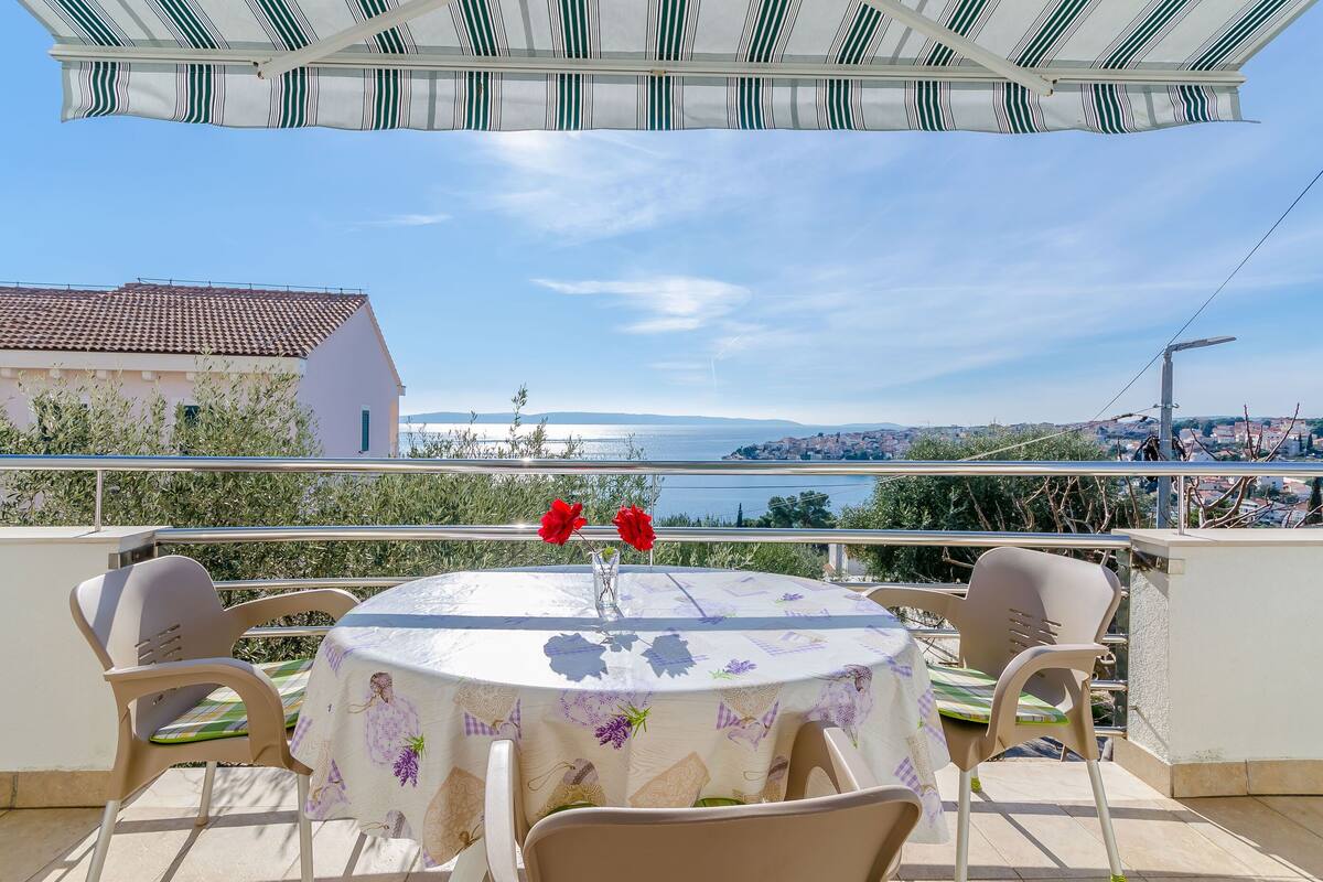 Une table ronde recouverte d’une nappe à motifs floraux, deux chaises beiges et un vase rempli de fleurs rouges, le tout sur un balcon donnant sur une vue sur la côte.