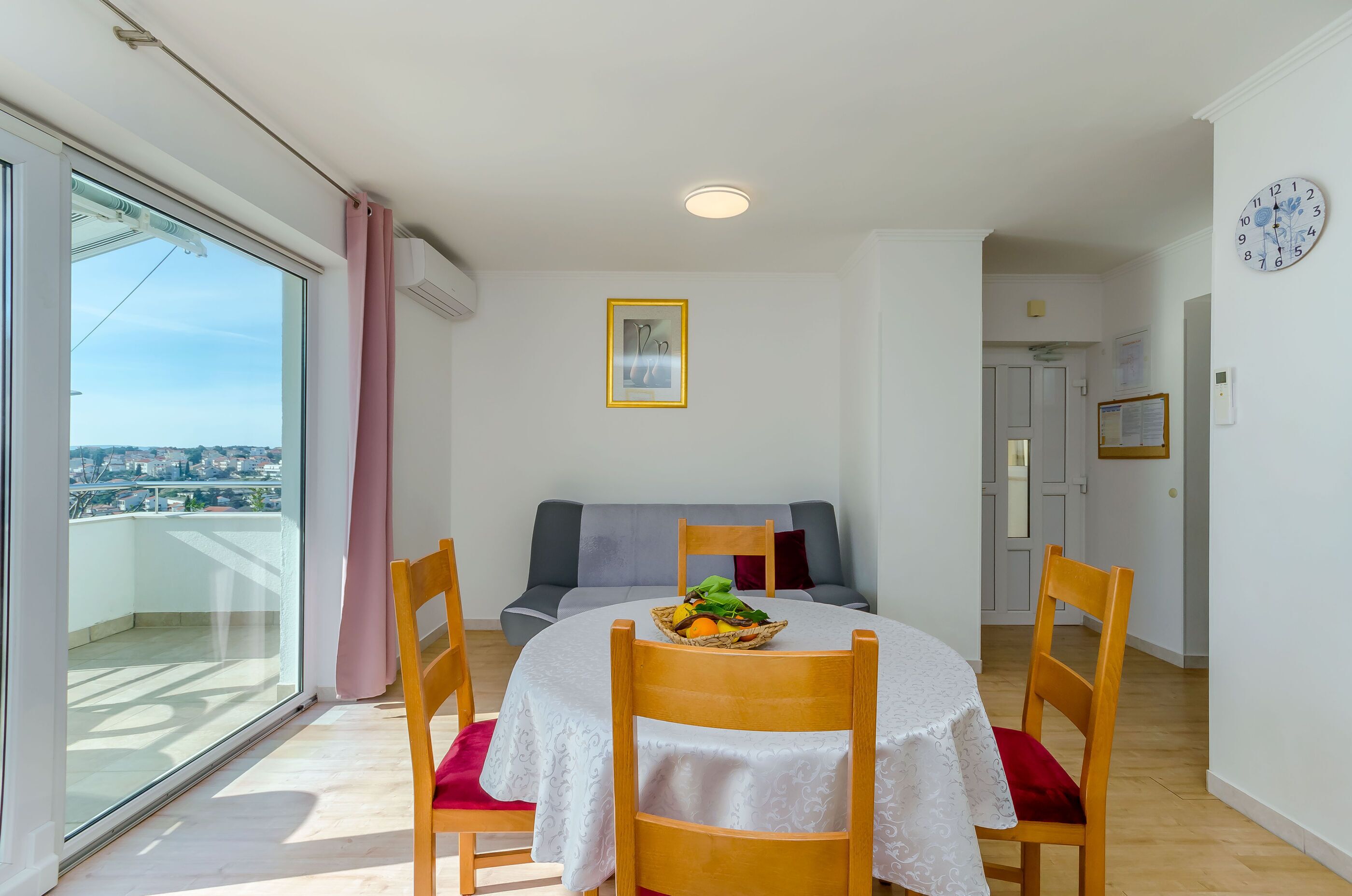 A dining area with a round table, chairs, and a fruit basket. There is a balcony with a view of the city.
