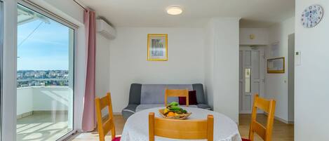 A dining area with a round table, chairs, and a fruit basket. There is a balcony with a view of the city.