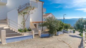 Ein modernes, weißes Gebäude mit einer Treppe, die zu einem Balkon führt, umgeben von Grün und mit freiem Blick aufs Meer.