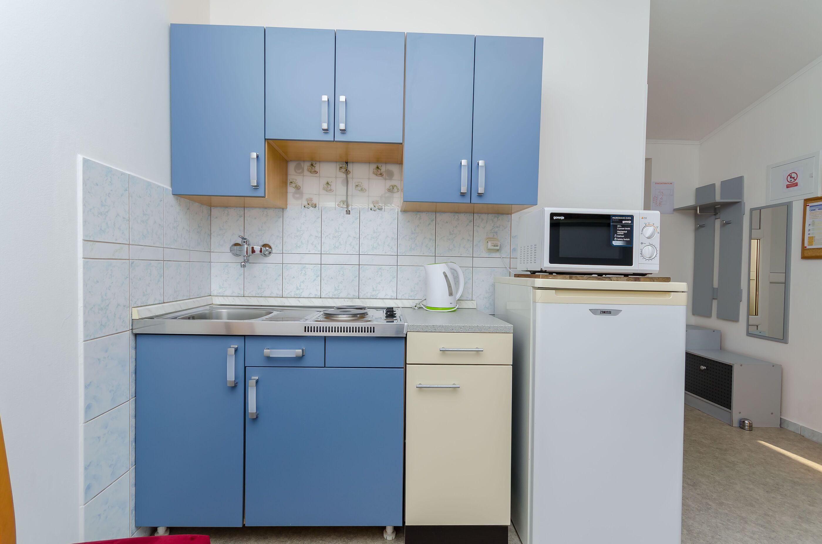 A compact kitchen with blue cabinets, a microwave, a kettle, and a small refrigerator.