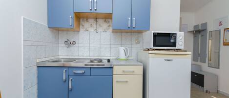 A compact kitchen with blue cabinets, a microwave, a kettle, and a small refrigerator.