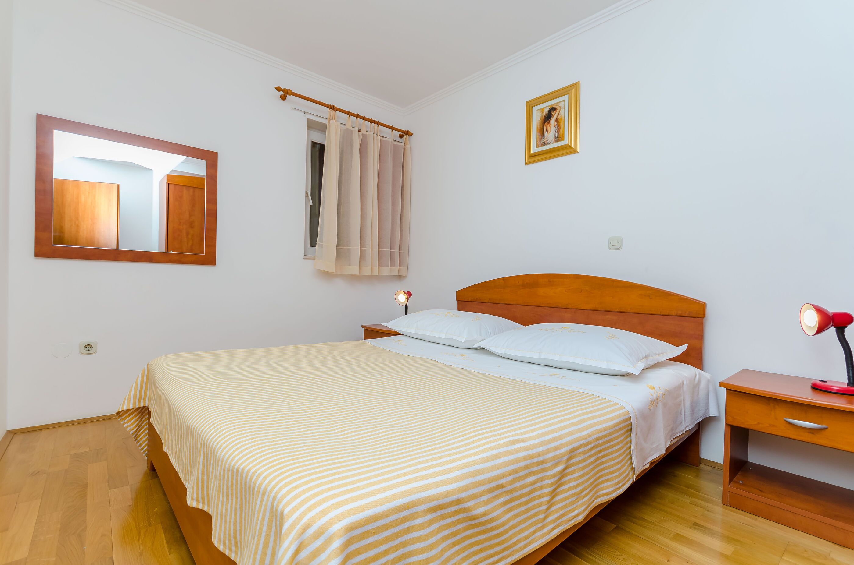 Una camera da letto con un letto in legno, un comodino con una lampada, uno specchio, una finestra con tende e un piccolo quadro appeso al muro.