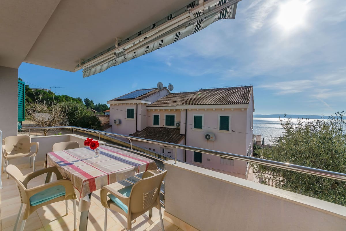 Un balcon avec une table et des chaises, donnant sur un immeuble avec un toit en tuiles et offrant une vue sur la mer.
