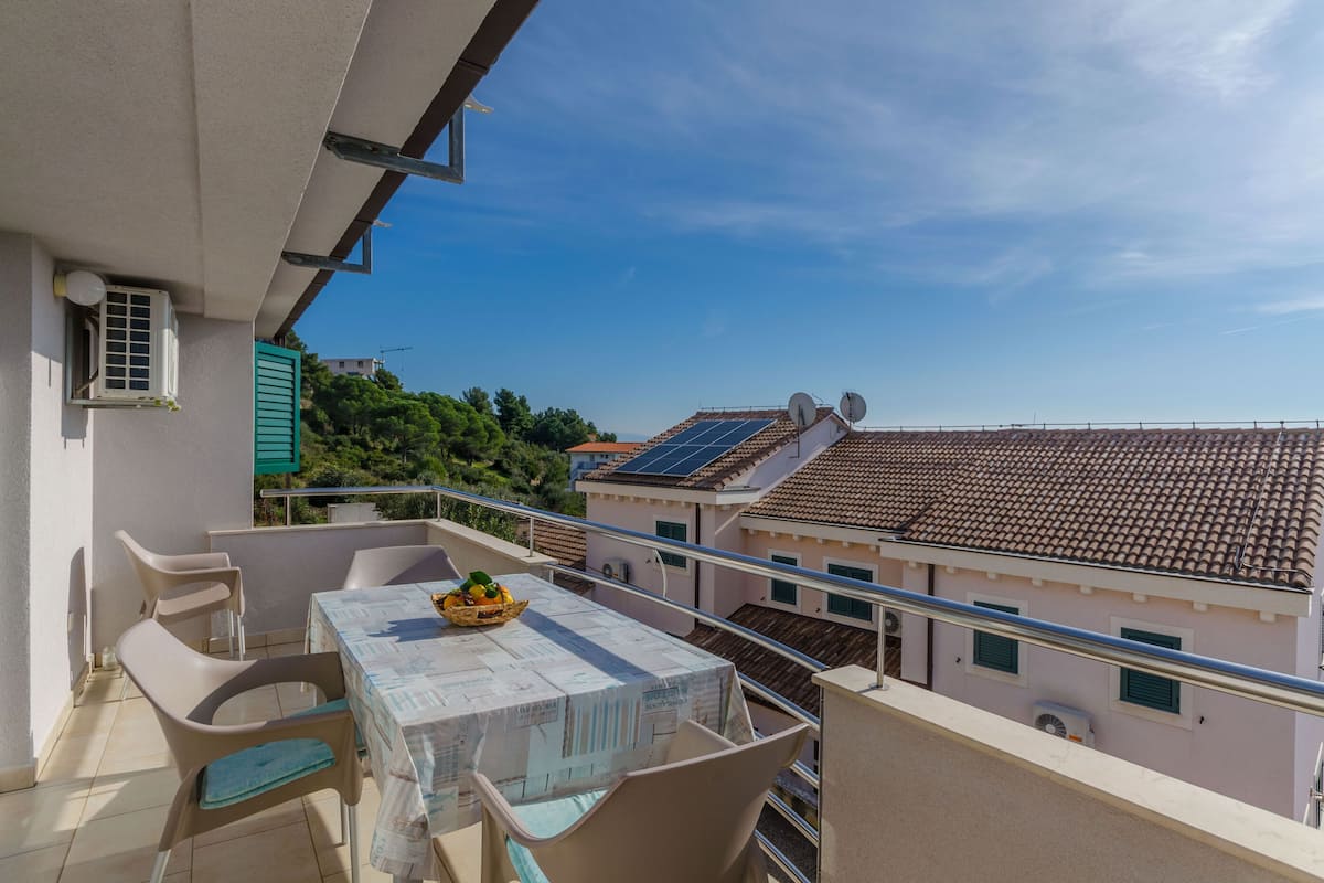 Un balcon avec une table et des chaises, un panier de fruits et une vue sur les bâtiments voisins, équipés de panneaux solaires.