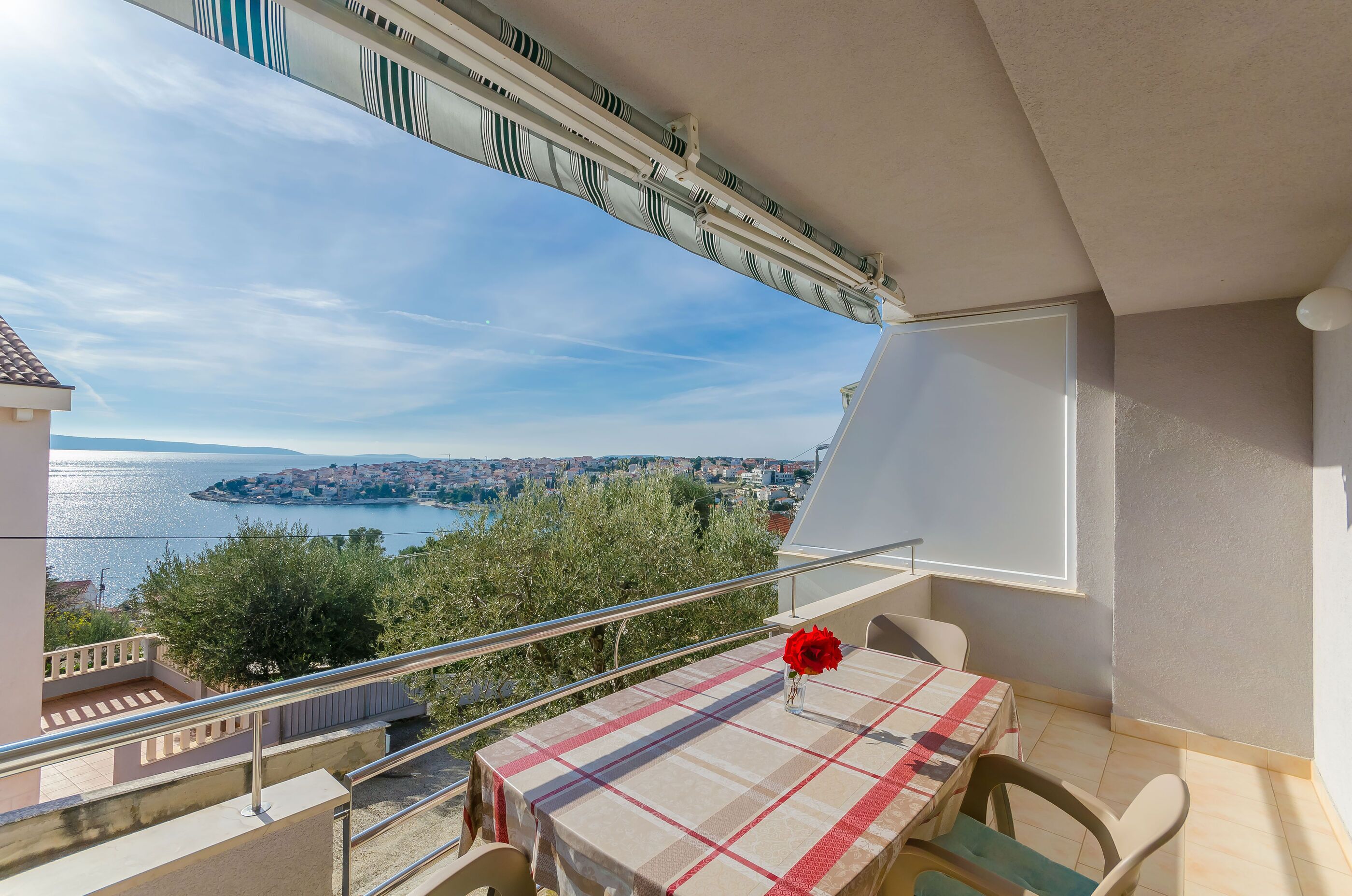 A balcony with a table and chairs, overlooking a coastal town and the sea.