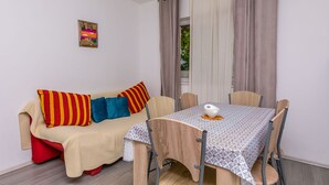 A room with a sofa, a dining table, and chairs. There is a window with curtains, a small painting on the wall, and a door leading outside. - Apartments KK (Omis)