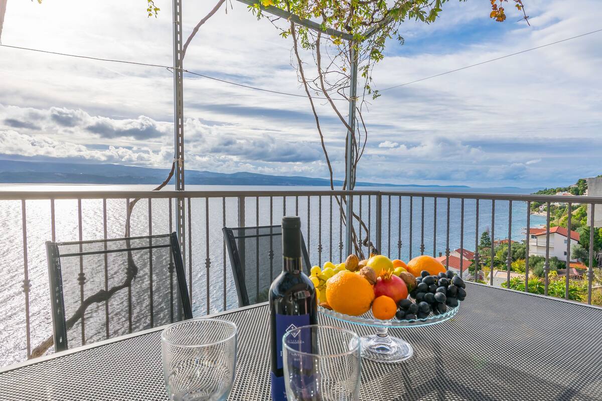 Un balcone con un tavolo apparecchiato per due, con una bottiglia di vino, una ciotola di frutta e due bicchieri.