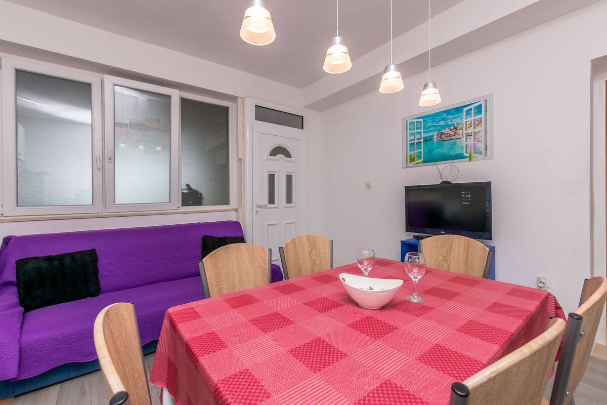 Una stanza con un divano viola, un tavolo a scacchi rossi, sedie di legno, una TV e un quadro raffigurante una vista mare.