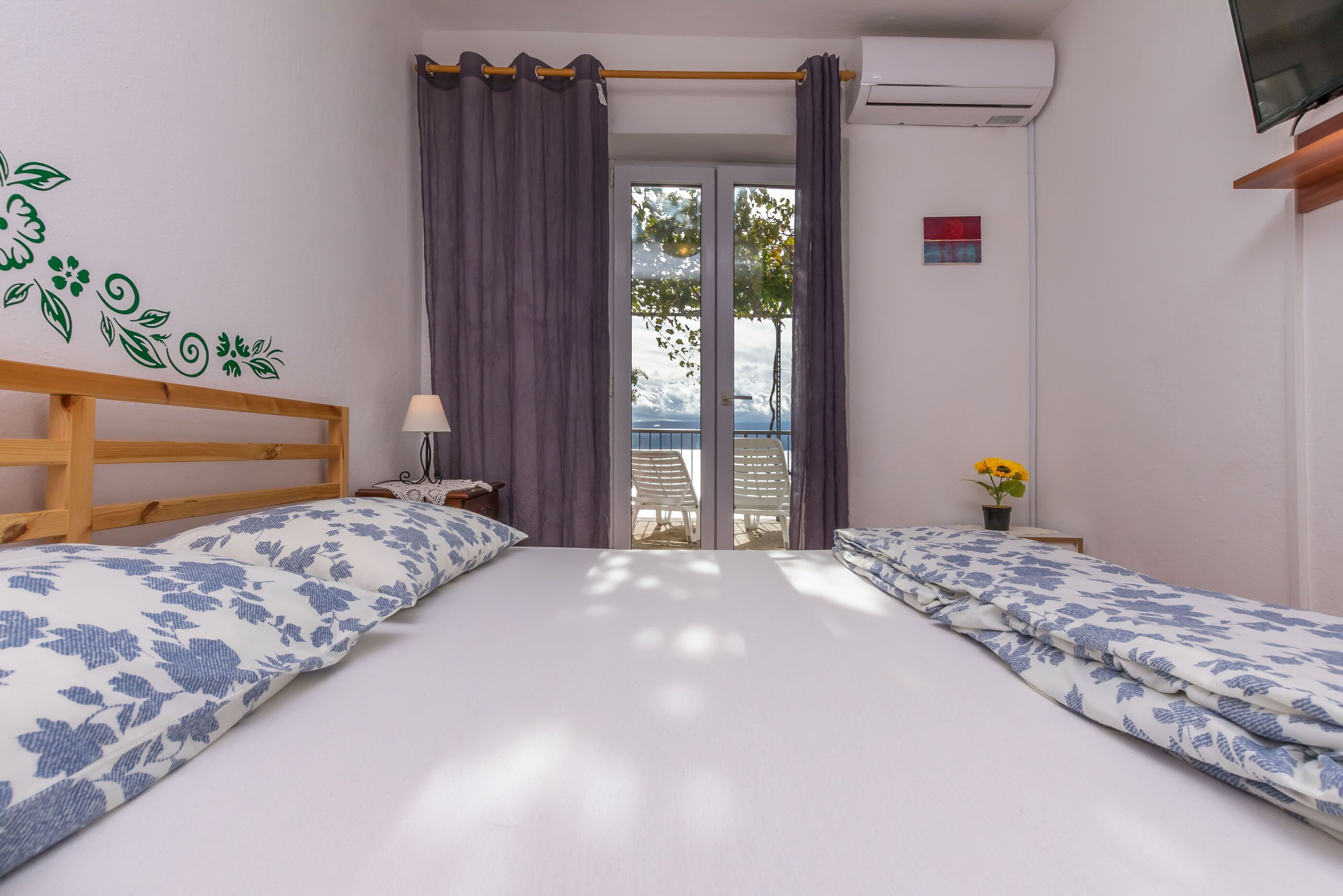 Quarto com duas camas, janela com cortinas, uma mesinha com abajur e vista para o exterior.