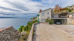 A coastal building with a stone terrace, a car parked under a covered area, and a view of the sea. - Apartments KK (Omis)