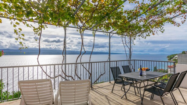 An outdoor terrace with a table and chairs, overlooking a body of water and a mountainous landscape. - Apartments KK (Omis)