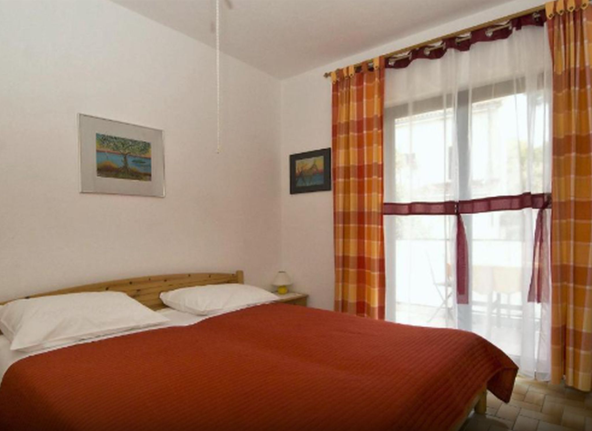 Une chambre à coucher avec un lit en bois, des oreillers blancs, une couverture rouge, une petite table de chevet avec une lampe, et deux tableaux accrochés au mur.