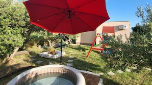 Un bain à remous avec un parasol rouge, entouré d’un jardin arboré et doté d’une terrasse en bois.