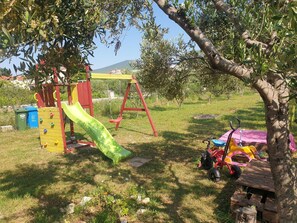 Une aire de jeux avec un toboggan, des balançoires et une petite table placée sous un arbre.
