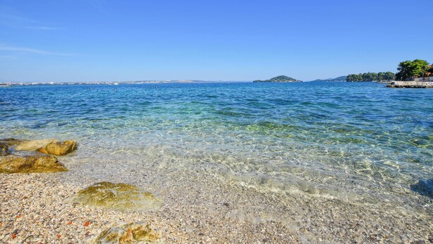 A clear view of a pebble beach with calm waters and a distant island. - Apartments Toni (Preko)