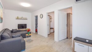 A modern living room with a grey sofa, a TV, and a small table with flowers. There is a wooden cabinet and a door leading to another room. - Apartments Toni (Preko)
