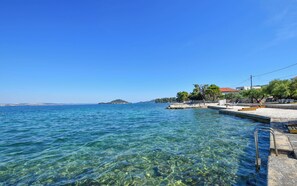 A clear coastal view with a pier extending into the sea, a small island in the distance, and a building with a red roof on the right. - Apartments Toni (Preko)