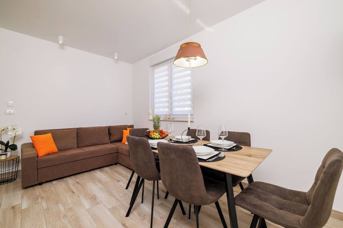 Ein modernes Wohnzimmer mit einem braunen Ecksofa, einem Holzesstisch mit vier Stühlen und einer hängenden Pendelleuchte.