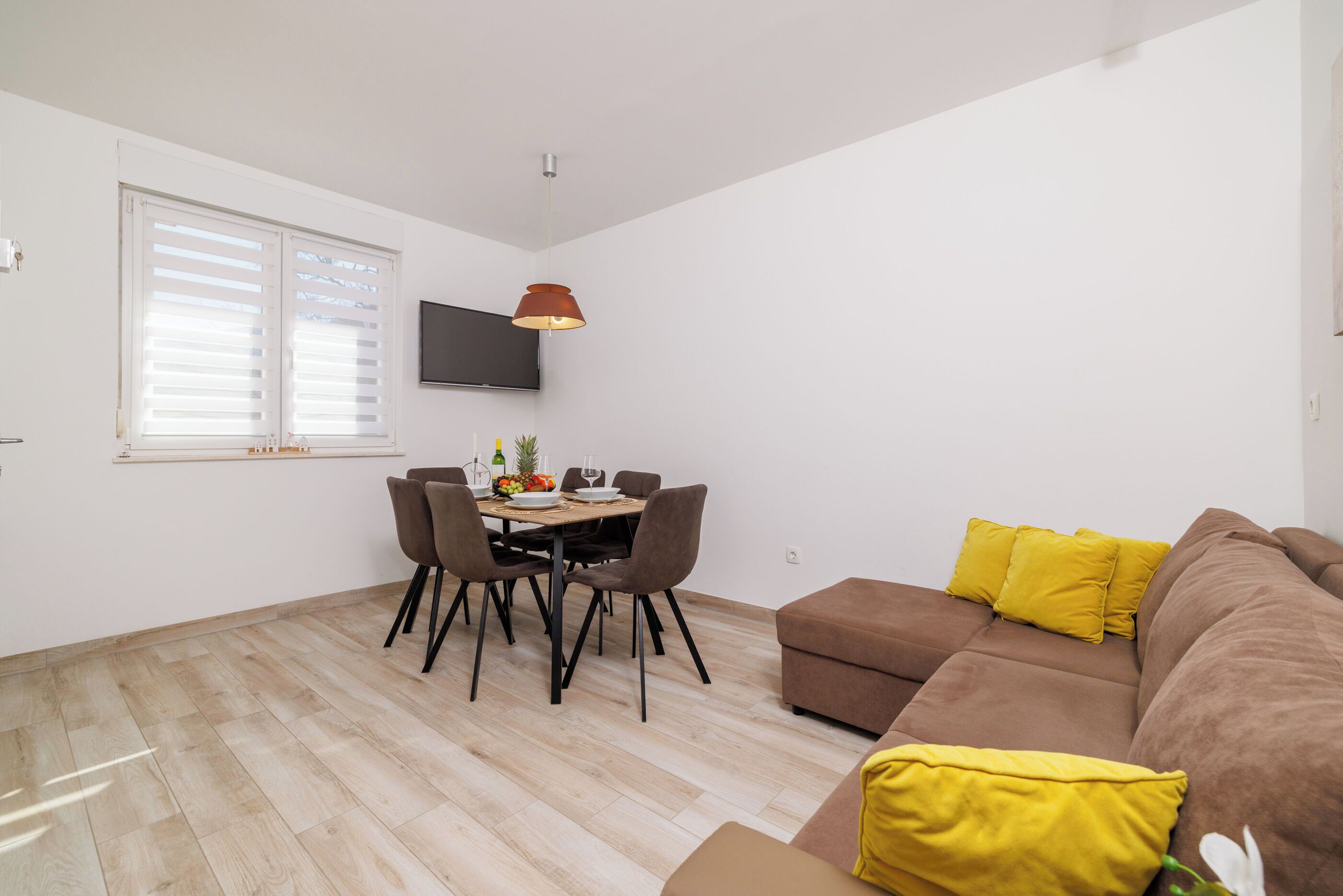 A modern living room with a brown sofa, yellow cushions, a dining table with chairs, and a flat-screen TV mounted on the wall.