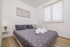A bedroom with a bed, pillows, a painting, a window, and a nightstand with a lamp. - Apartments Margita 2 (Vinodol)
