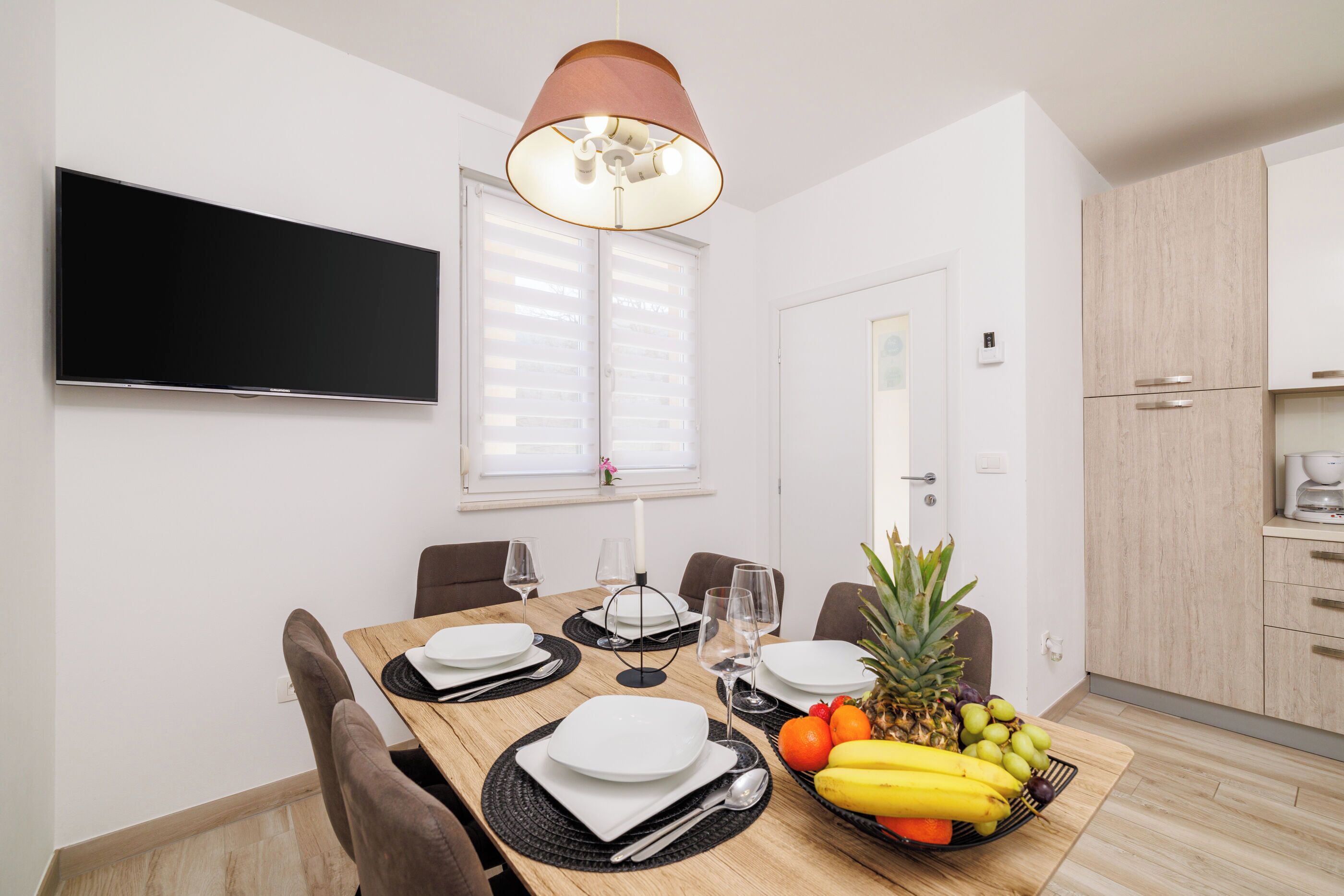 A dining area with a table set for four, a fruit bowl, and a television mounted on the wall.