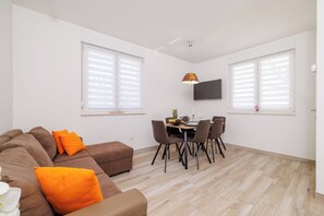 Ein modernes Wohnzimmer mit einem braunen Sofa, orangefarbenen Kissen, einem Esstisch mit Stühlen und einem an der Wand befestigten Flachbildfernseher.
