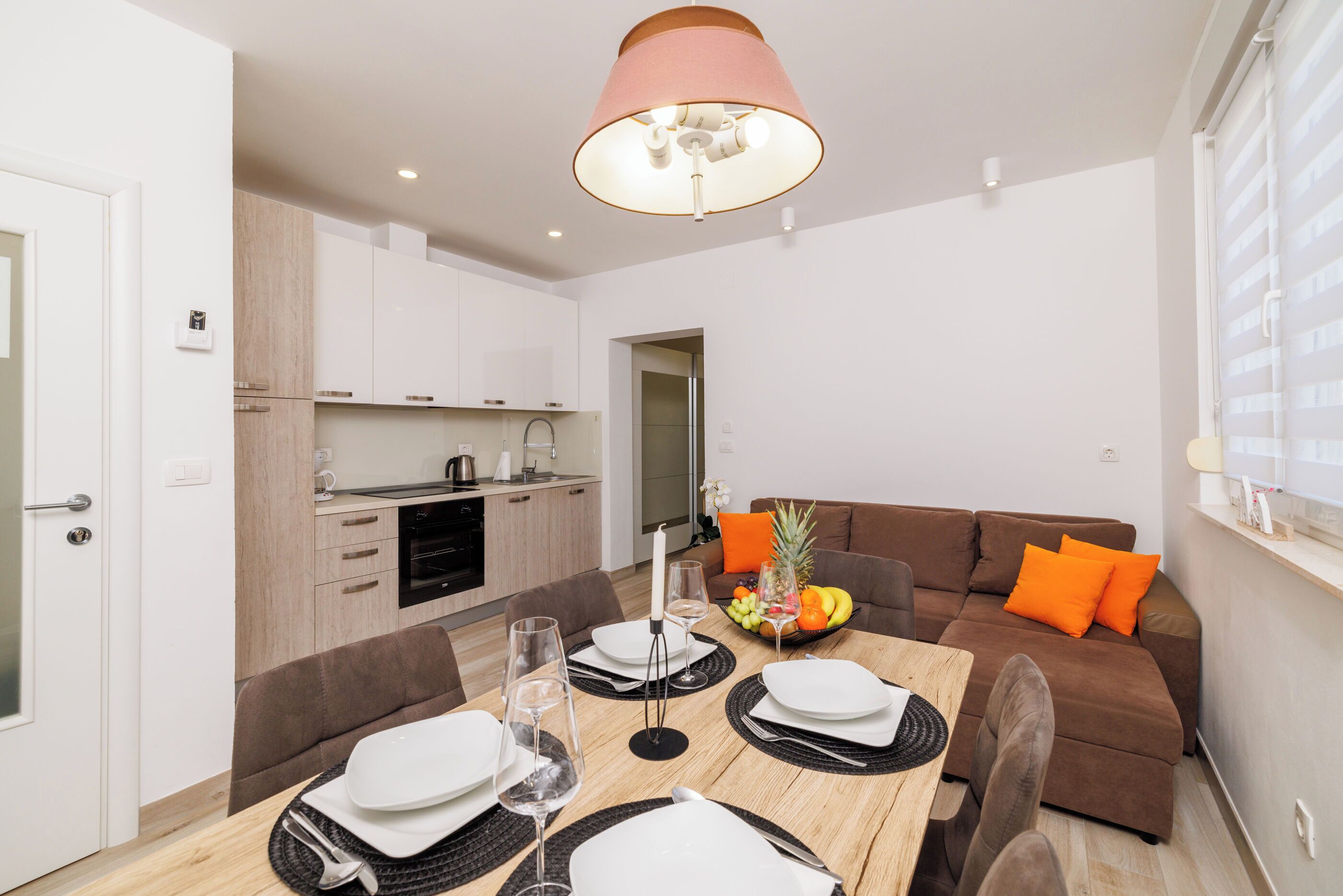 A modern dining area with a wooden table set for four, a brown sofa with orange cushions, and a kitchen in the background.