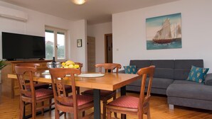 Een eetruimte met een houten tafel, stoelen en een schaal fruit. Links staat een televisie en aan de muur hangt een schilderij.