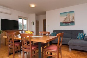 A dining area with a wooden table, chairs, and a fruit bowl. There is a television on the left and a painting on the wall. - Apartments La Mer (Pasman)