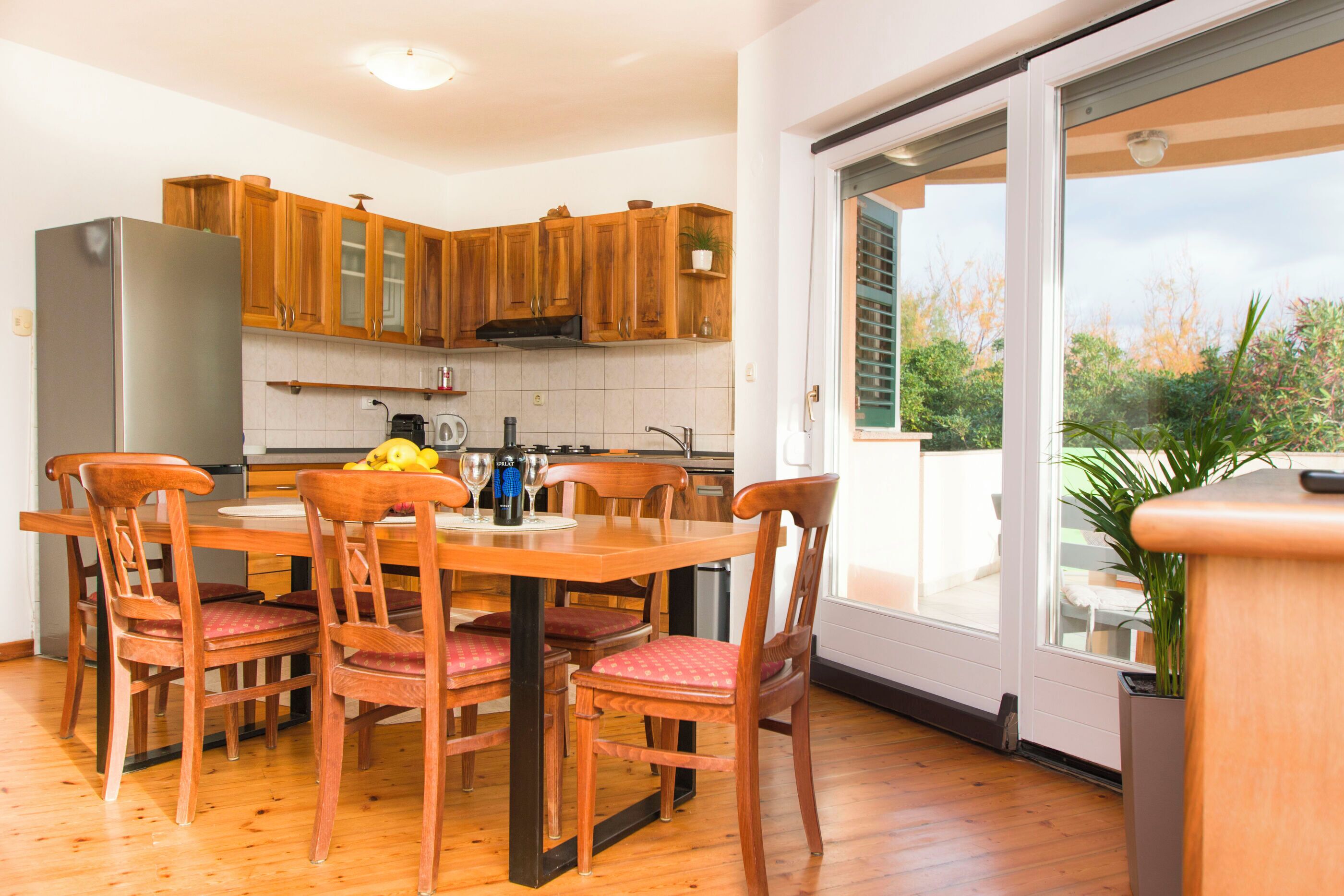 A modern kitchen with wooden cabinets, a dining table with chairs, and a view through a sliding glass door.