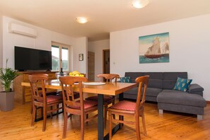 A dining area with wooden chairs and a table, a gray sofa, a painting of a sailboat, and a television on the wall. - Apartments La Mer (Pasman)