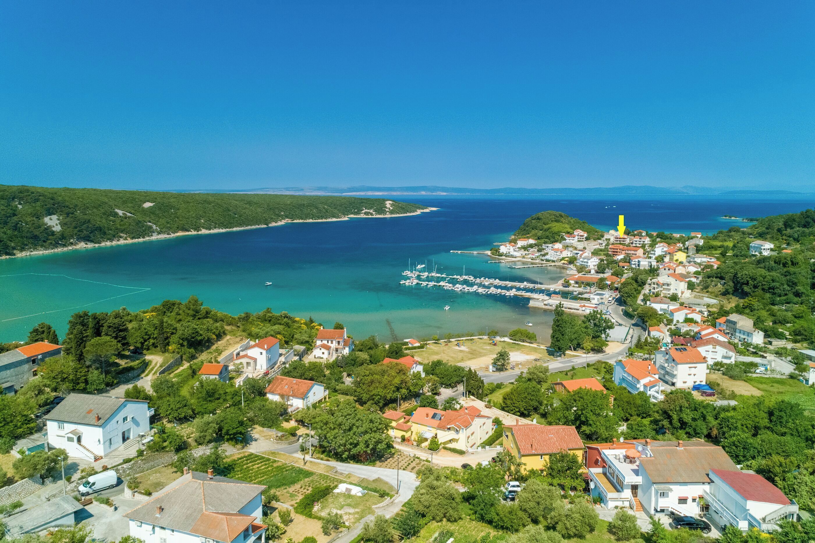 An aerial view of a coastal town with houses, a marina, and a clear blue sea.